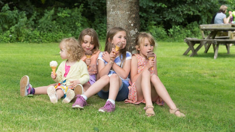 Four young girls sat down on the grass leaning on a tree, each eating an ice cream in a cone.